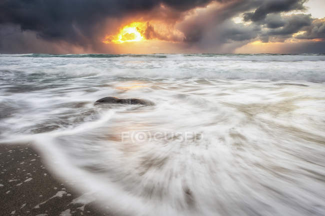 Flou de la marée se jetant sur le rivage sablonneux le long de la côte et un soleil doré culminant à travers les nuages orageux ; Nordland, Norvège — Photo de stock