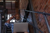 Senior man talking on mobile phone while using laptop on staircase — Stock Photo