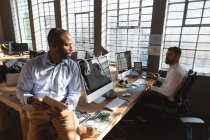 Front view close up of a young African American man sitting on a desk using a tablet computer and talking with a young Caucasian male colleague sitting at a desk using a computer by a window in a creative office — Stock Photo