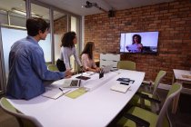 Side view of a young mixed race woman and a young Caucasian woman and man working in the office of a creative business, in a meeting with a female colleague communicating by video link, visible on a wall mounted screen — Stock Photo