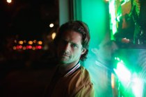 Portrait of a young Caucasian man looking to camera in the evening with green neon light from a shop window behind him — Stock Photo