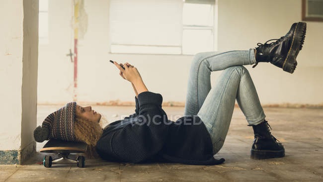 Side view of a young Caucasian woman with curly hair wearing a black shirt and beanie lying on the floor with head on a skateboard while using a mobile phone — Stock Photo