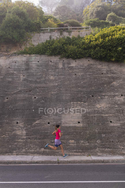 Side view of a young Caucasian woman wearing sports clothes running past a high retaining wall during a workout on a sunny day — Stock Photo