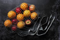High angle close up of red apples and oranges in grey net bag on black background. — Stock Photo