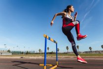 Strong young woman in sportswear leaping over hurdle against blue sky during workout on stadium — Stock Photo