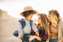 Cheerful mother in checkered shirt holding casual toddler as joyful female friend putting hat on child on nature at daytime — Stock Photo