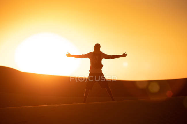 Back view of unrecognizable tourist with outstretched arms standing against bright cloudless sundown sky in desert — Stock Photo