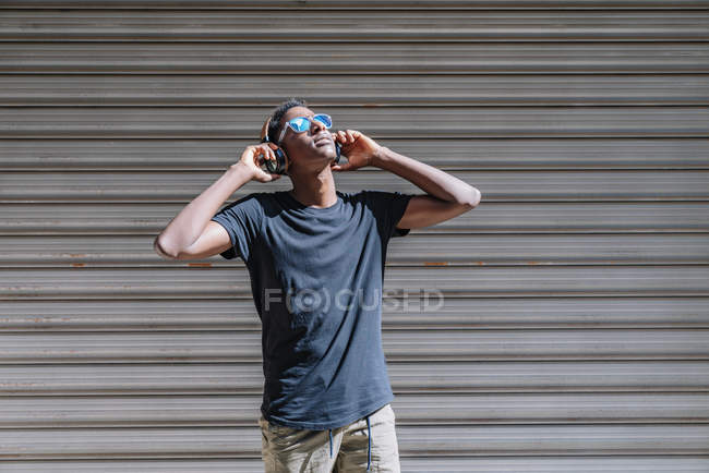 Stylish modern African American man in sunglasses and headphones listening to music at striped wall in street — Stock Photo