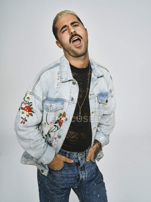 Young ethnic man making grimace face looking at camera wearing trendy denim jacket with floral pattern while standing against gray background — Stock Photo