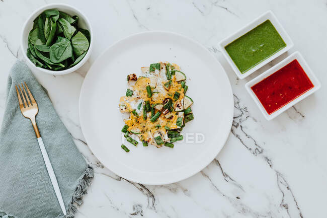 De cima prato com abobrinha e salada de feijão verde com queijo colocado na mesa de mármore perto tigela com folhas de espinafre fresco e molhos de abacate e tomate ao lado de guardanapo com garfo — Fotografia de Stock