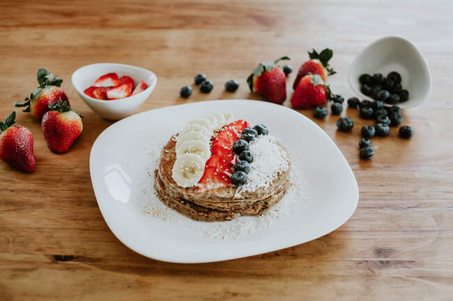 Pilha de panquecas saborosas servidas em prato com pedaços de banana e morango e mirtilos frescos com flocos de coco durante o café da manhã — Fotografia de Stock