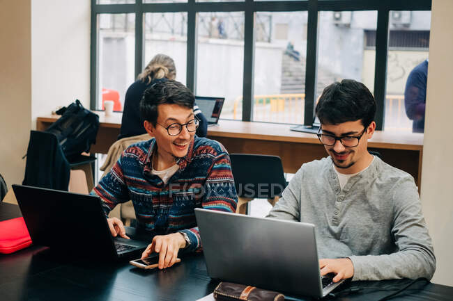 Positive coworkers in casual clothes and eyeglasses sitting at desk and working on project on laptops during workday in modern office — Stock Photo