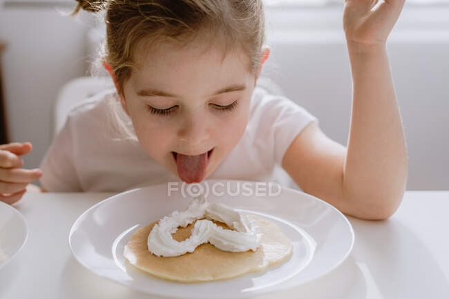 Thrilled little child in casual t shirt sitting at table with plate of tasty pancake garnished with heart shaped whipped cream — Stock Photo