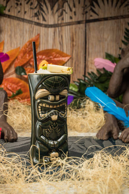 Large sculptural tiki cup filled with booze decorated with straw and fruits placed on green rug against dry grass — Stock Photo