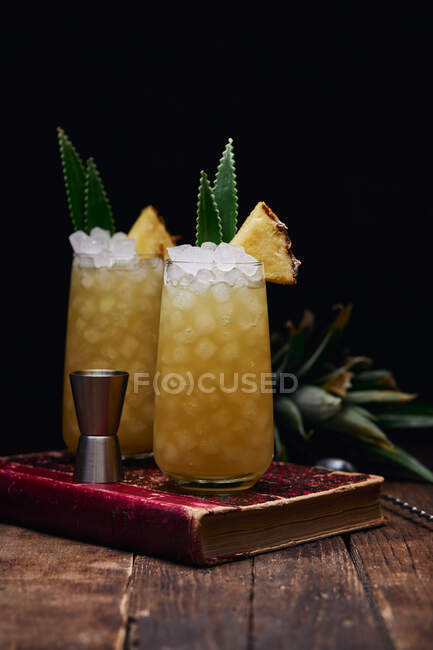 Wooden table with glasses of fresh yellow cocktails with ice cubes and pineapple pieces and leaves near spoon and shot glass placed on red book on black background — Stock Photo