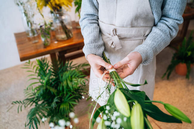 Cultivé fleuriste femelle méconnaissable en vêtements décontractés et tablier attachant bouquet délicat de lys orange et gypsophila blanc dans la boutique de fleurs — Photo de stock