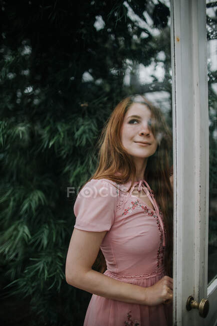 Dreamy positive redhead female in dress standing near glass doorway of glasshouse with lush plants — Stock Photo