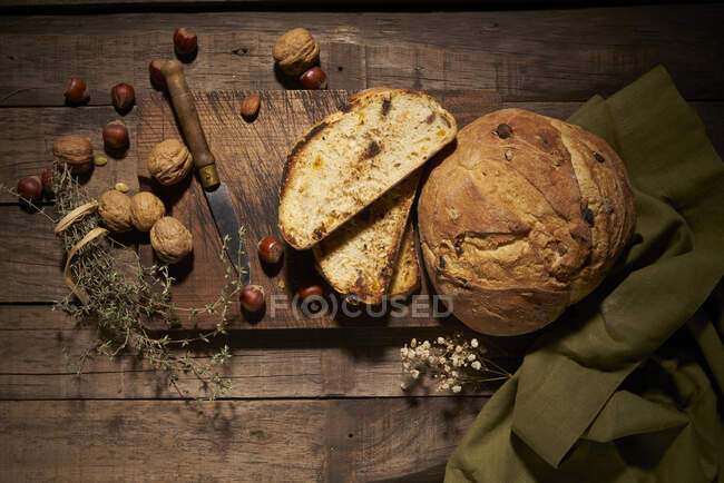 Top view of delicious homemade sourdough bread loaf with nuts placed on wooden board with knife and aromatic herbs on rustic table — Stock Photo