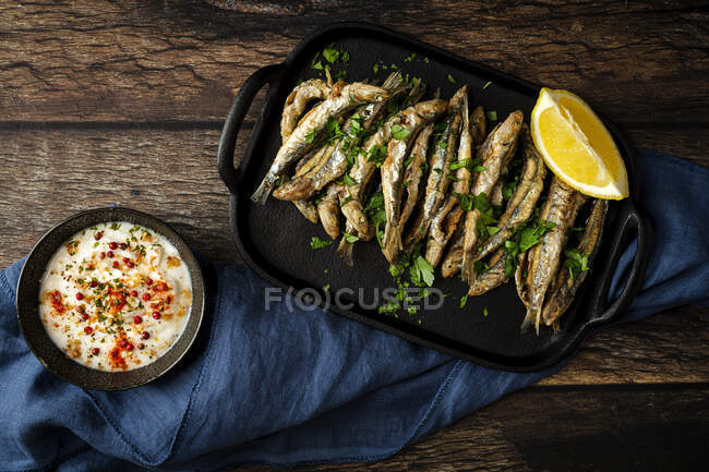 Top view of tasty fried anchovies with juicy lemon piece and chopped parsley on tray against savory sauce — Stock Photo