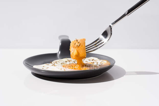 Metal fork with fresh piece of bread dipped in liquid yolk of fried egg served on skillet on white background — Stock Photo